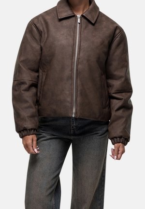 Personne portant une veste marron zippée avec poignets et col élastiques, associée à un jean noir délavé sur un fond clair uni.