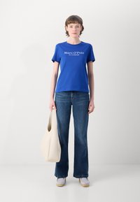 Královsky modré tričko s bílým logo textem, kombinované s tmavě modrými širokými džíny a off-white taškou. Model stojí proti neutrálnímu pozadí.