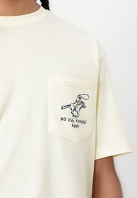 T-shirt en coton jaune clair avec une poche sur la poitrine, présentant un graphique bleu d'un cowboy sur un cheval et un texte en dessous disant "NE SERT À RIEN DE FUIR."