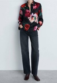 Blouse noire à fleurs rouges, roses et vertes, à manches longues et col, associée à un jean droit foncé et des chaussures marron foncé.