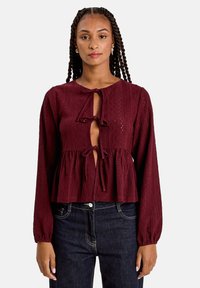 Blouse bordeaux à manches longues, présentant un motif texturé, une fermeture à lacer à l'avant et un design à taille peplum. Assortie à un jean foncé.