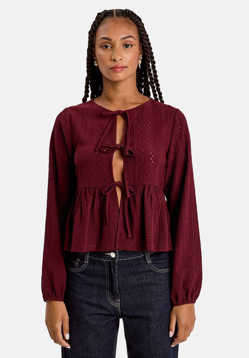 Blouse bordeaux à manches longues, présentant un motif texturé, une fermeture à lacer à l'avant et un design à taille peplum. Assortie à un jean foncé.