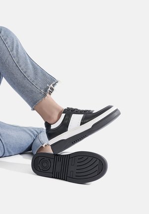 Sneaker bianco e nero con una tomaia in pelle liscia, dettagli perforati, una robusta intersuola bianca e una suola in gomma nera testurizzata.