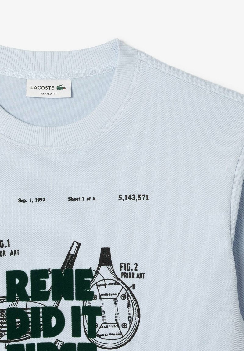 Chemise courte à manches courtes bleu clair avec un tissage texturé, col rond et impression graphique verte avec des illustrations de raquettes de tennis et des détails en vernis.