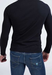 Uomo che indossa una maglietta nera a maniche lunghe aderente e jeans di denim blu, mostrato di spalle su uno sfondo bianco semplice.