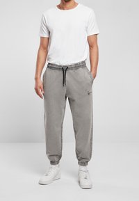 Šedé tepláky s elastickými manžetami a stahovací šňůrkou v pase, s bočními kapsami a malým černým logem na levém stehně.