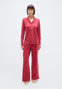 Femme debout pieds nus portant un pyjama à manches longues rouge avec des boutons, orné d'un motif de petits cœurs blancs et d'un liseré blanc sur le col et les poignets.