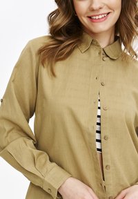 Khaki koszula zapinana na guziki z kołnierzykiem, długimi rękawami i drewnianymi guzikami. Posiada luźny krój i jest noszona na paskowanym topie.