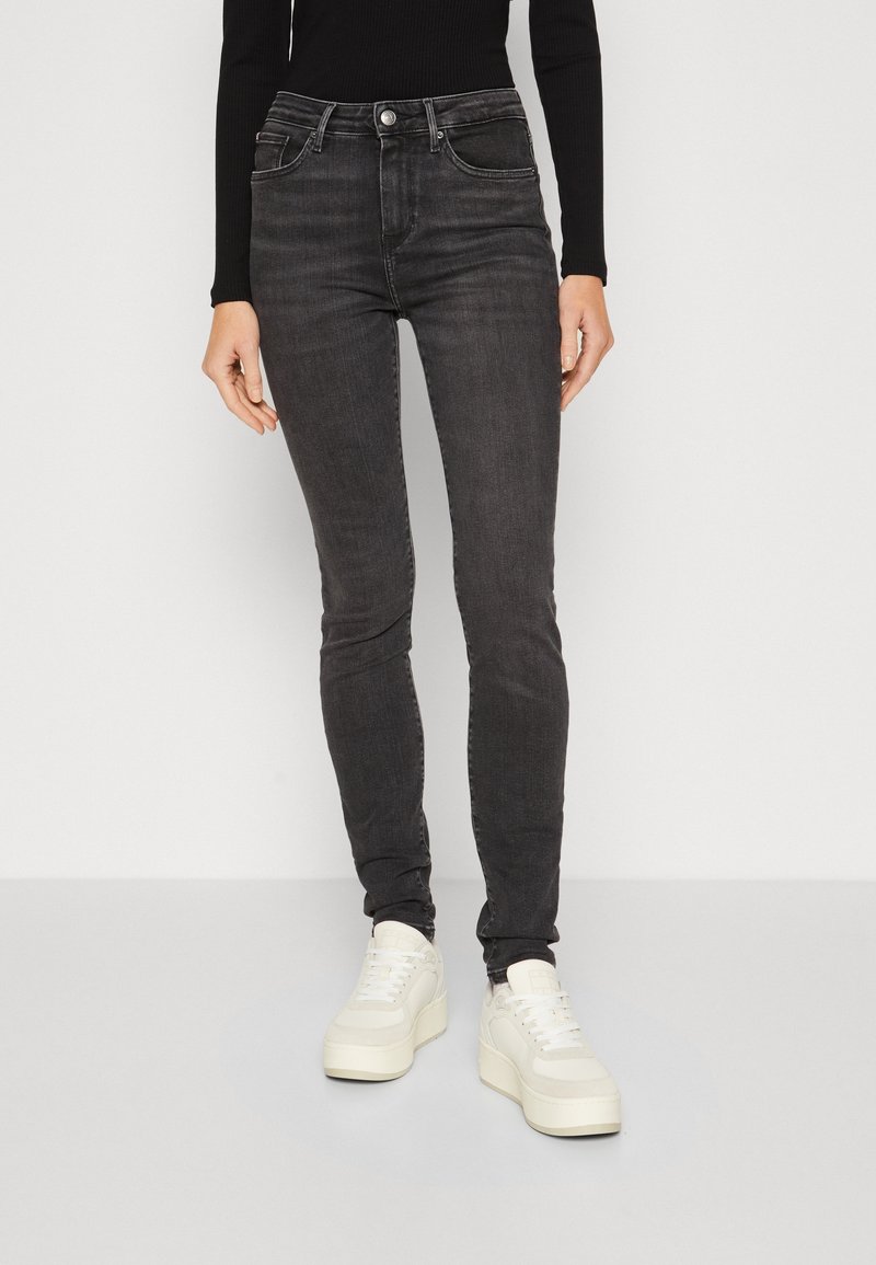 Jeans negros ajustados de denim con cintura alta, dos bolsillos frontales y costuras sutiles. Combinados con zapatillas blancas de plataforma.