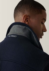 Abrigo de lana azul marino con un alto cuello, con un interior gris y texto bordado "SOY CONNABLE" en el cuello, acabado elegante y texturizado.