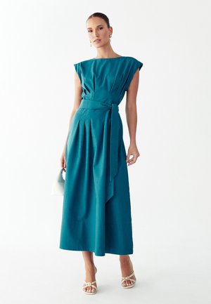 Kobieta w turkusowej, bezrękawowej sukience midi z paskiem i plisami, trzymająca małą białą torebkę, ubrana w beżowe buty na obcasie z odkrytymi palcami.