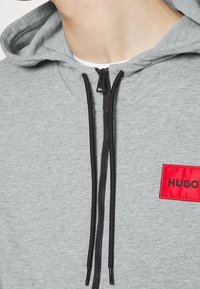 Felpa grigia con cappuccio e cordini neri, chiusura con zip e un patch rettangolare rosso con la scritta "HUGO" in testo nero. Tessuto morbido, design casual.