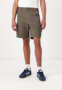 Shorts cargo vert olive avec deux poches latérales, fermeture à bouton et coupe droite. Portés avec des baskets marines et des chaussettes blanches.