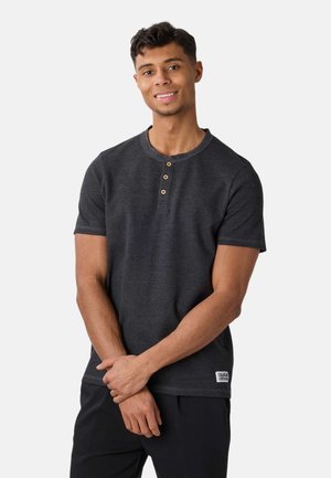 Giovane sorridente che indossa una maglietta henley a maniche corte grigio scuro con tre bottoni e pantaloni neri, in piedi davanti a uno sfondo semplice.