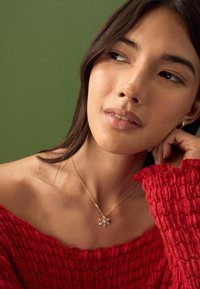 Gold necklace with floral pendant, red textured off-the-shoulder top, skin tone visible, subtle makeup, green background.