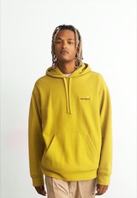 Haut en molleton jaune avec une poche kangourou à l'avant, capuche avec cordon de serrage et logo brodé. Le modèle porte un pantalon beige. Design simple.