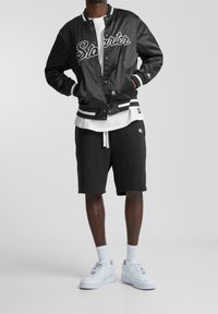 Veste bomber en satin noir avec inscription "Starter", bordure rayée blanche et noire. Portée avec un t-shirt blanc et un short noir, assortie à des baskets blanches.