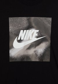 Juodas marškinėlis su dideliu sidabriniu piešiniu, kuriame yra balta Nike logotipas, ant tekstūruoto metalinio fono.