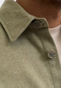 Olivengrønn skjorte med krage og teksturert stoff, med synlig knapp og pent sydd kant. Nærbilde av krageområdet.