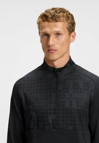 Top zip-up sportivo nero con un motivo a griglia testurizzato nella parte superiore, colletto alto e tessuto liscio nella parte inferiore, dotato di una cerniera centrale.