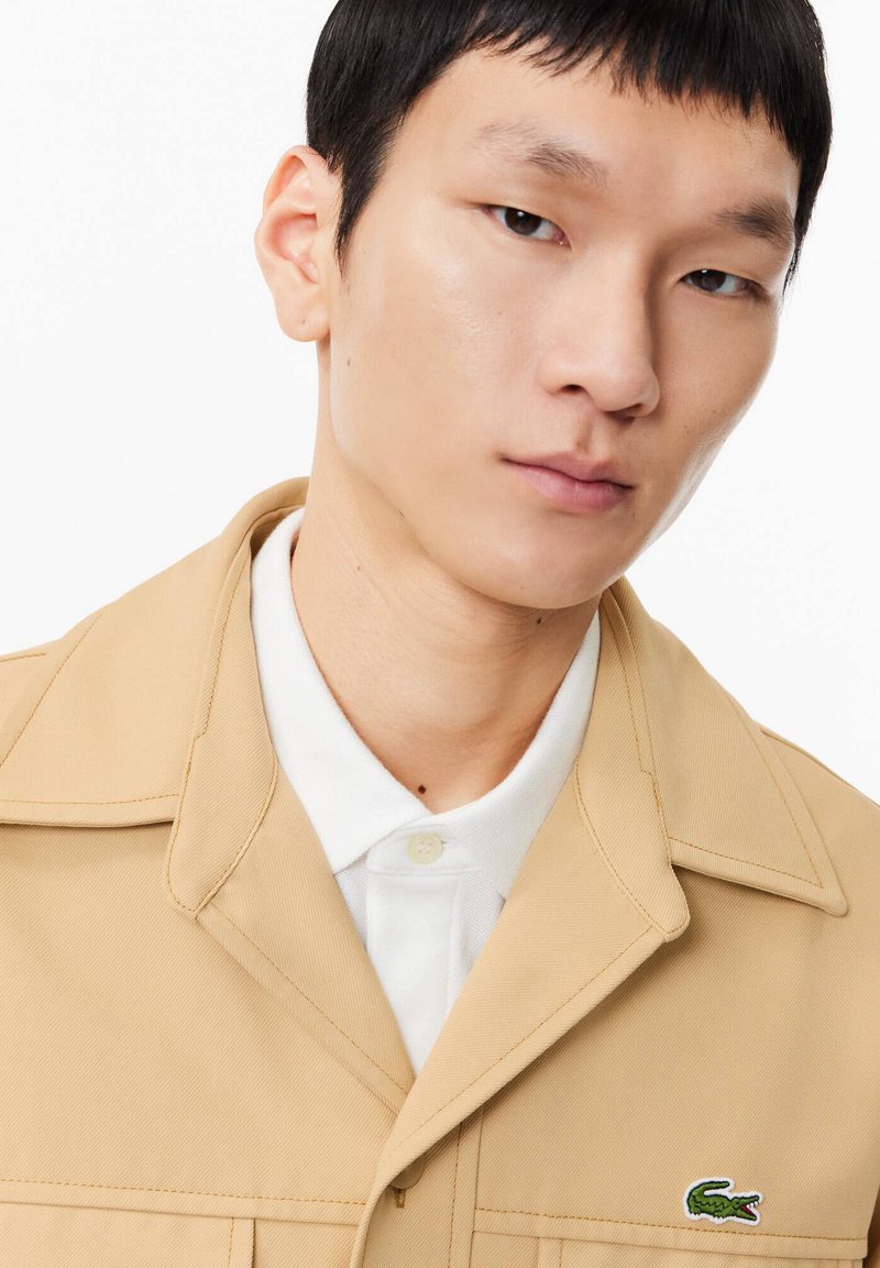 Beige Jacke mit strukturiertem Design und Umlegekragen, getragen über einem weißen Poloshirt; verfügt über einen kleinen grünen Logopatch auf der Brust.