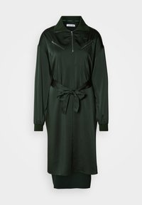 Σκούρο πράσινο φόρεμα με φερμουάρ, φτιαγμένο από λείο, γυαλιστερό ύφασμα. Διαθέτει γιακά, μακριά μανίκια και ζώνη στη μέση. Περιλαμβάνει τσέπες με φερμουάρ.