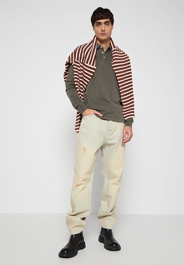 Khaki Langarm-Polo, hellblaue, verwaschene Jeans und schwarze Stiefeletten. Gestreifter braun-weißer Pullover über die Schultern gelegt.