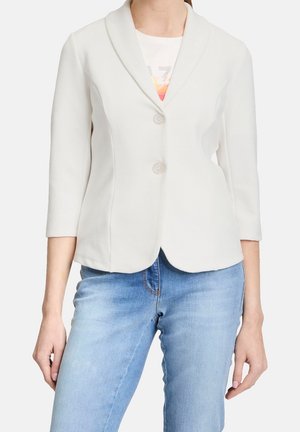 Donna che indossa un blazer bianco a due bottoni con maniche a tre quarti sopra una t-shirt grafica, abbinato a jeans in denim azzurro chiaro.