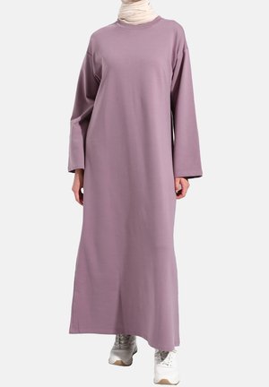 Femme portant une robe longue mauve ample à manches longues, des baskets blanches et une écharpe beige autour du cou sur un fond uni.