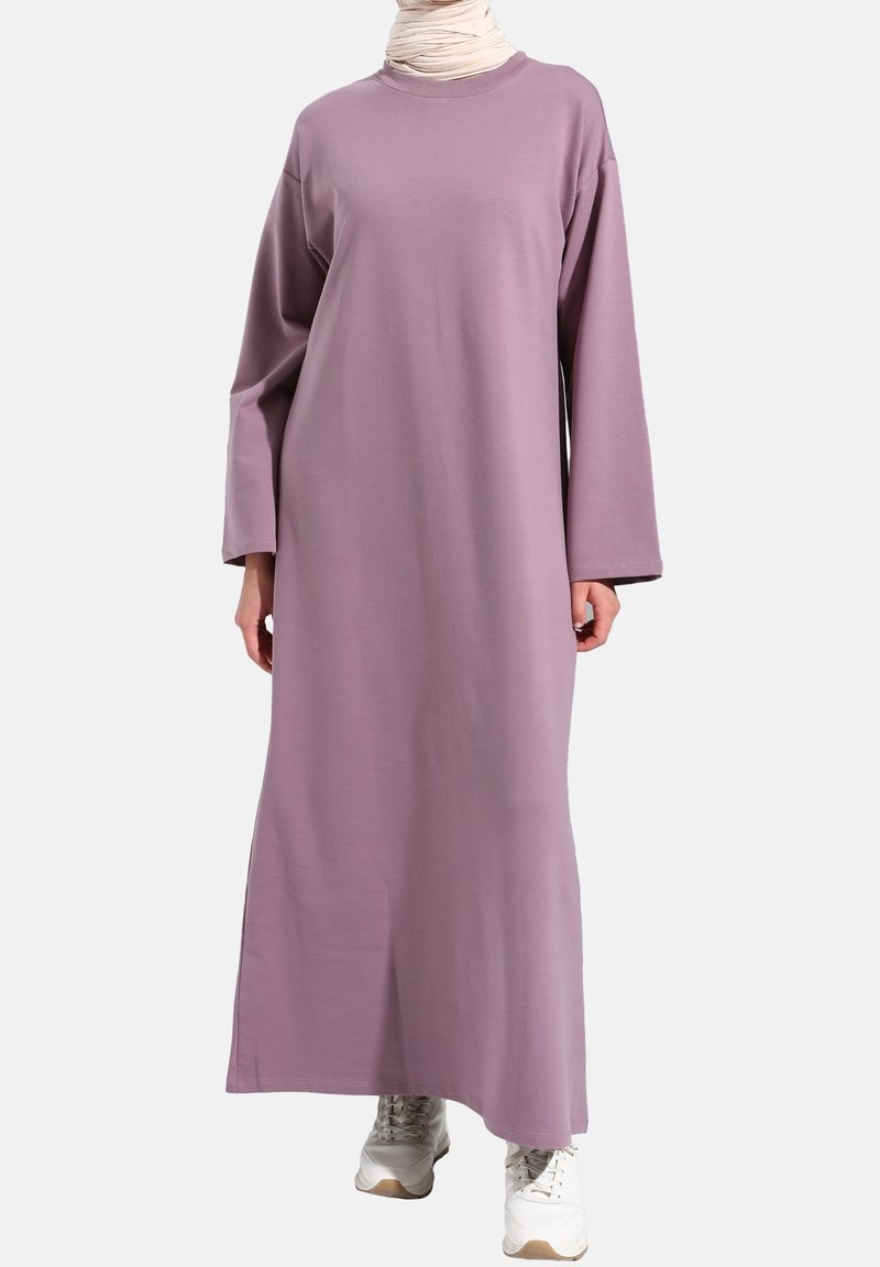 Femme portant une robe longue mauve ample à manches longues, des baskets blanches et une écharpe beige autour du cou sur un fond uni.