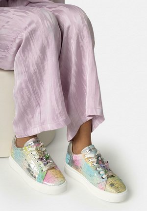 Persona vistiendo zapatillas de patrón de cocodrilo en colores pastel con decoraciones de joyas y detalles metálicos, combinadas con pantalones lavanda sueltos.