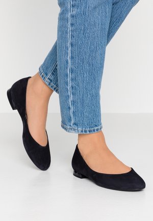 Bailarinas negras de ante con un tacón bajo, punta redonda y cortes laterales, combinadas con unos vaqueros azul claro con dobladillo. Textura suave y diseño minimalista.