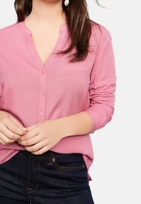 Camicia rosa con bottoni e maniche lunghe, realizzata in tessuto leggero. Presenta un colletto e una piccola tasca sul lato sinistro. Indossata con jeans scuri.