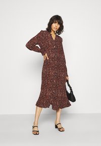 Robe longue à boutons, avec un motif floral rouge ; possède un ourlet évasé et des manches longues. Associée à un sac à main noir et des sandales noires à lanières.
