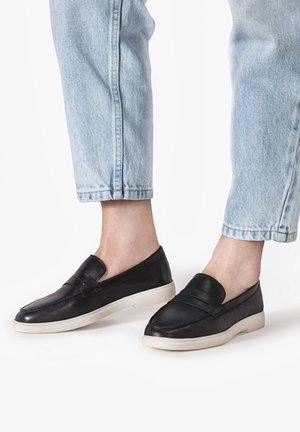 Zapatos negros de cuero sin cordones con un acabado liso, que presentan un diseño sutil de tira frontal y una suela exterior de goma blanca contrastante.