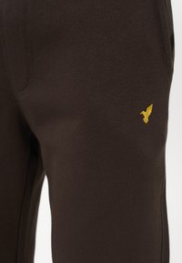 Pantalon marron foncé avec un petit logo d’oiseau jaune brodé près de la poche sur le haut de la cuisse.