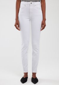Jeans blancos de talle alto con un corte ajustado, cinco bolsillos, herrajes plateados y una textura sutil, combinados con zapatos negros tipo slip-on.