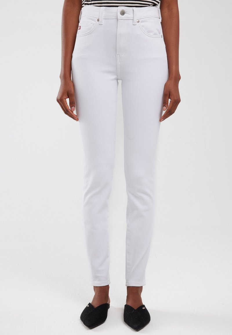 Jeans blancos de talle alto con un corte ajustado, cinco bolsillos, herrajes plateados y una textura sutil, combinados con zapatos negros tipo slip-on.