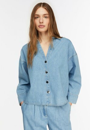 Junge Frau mit langen Haaren, die ein hellblaues Jeanshemd mit Knöpfen und passende Jeanshosen trägt, die Hände in den Taschen.