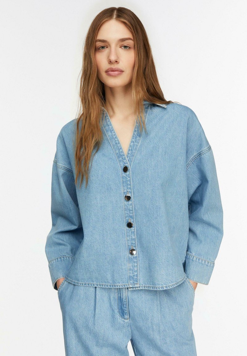 Junge Frau mit langen Haaren, die ein hellblaues Jeanshemd mit Knöpfen und passende Jeanshosen trägt, die Hände in den Taschen.