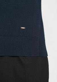 Pull en maille bleu marine avec ourlet et poignets côtelés, comportant une petite étiquette logo argentée. Texture lisse et coupe ajustée.