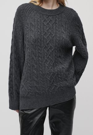 Femme portant un pull torsadé gris foncé et un pantalon en cuir noir, debout avec une main dans le dos.