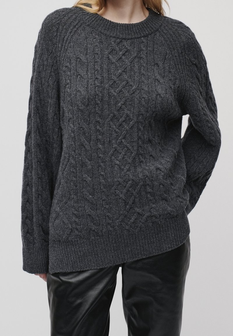 Femme portant un pull torsadé gris foncé et un pantalon en cuir noir, debout avec une main dans le dos.