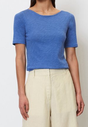 Kvinde iført en tætsiddende blå bluse med korte ærmer, stoppet ned i højtaljede, cremegule bukser med brede ben, stående mod en hvid baggrund.