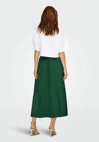 Top bianco corto a maniche corte abbinato a una gonna maxi verde fluida e plissettata. Tacchi metallici con cinturini visibili.