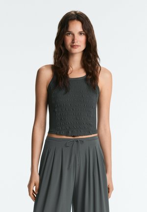 Femme aux cheveux bruns ondulés portant un crop top sans manches texturé gris foncé et un pantalon ample gris foncé avec une taille à cordon.