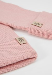 Rosa gestrickte Pulloverärmel mit gerippten Bündchen, versehen mit einem Stoffetikett, auf dem ein Logo an der Seite gestickt ist. Weiche Textur und glatte Oberfläche.