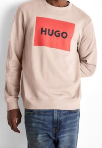 Sudadera de felpa en rosa claro con un gráfico rectangular rojo que presenta "HUGO" en letras negras en negrita. Puños y dobladillo acanalados.
