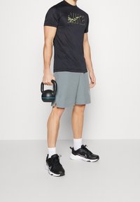 Camiseta deportiva negra con logotipo de camuflaje, pantalones cortos grises y zapatillas negras. Sosteniendo una kettlebell negra con acentos azules.