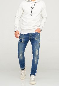 Hellgrau Hoodie mit schwarzen Kordeln, kombiniert mit durchgewetzten blauen Jeans mit Rissen und Farbspritzern, getragen mit beigen Turnschuhen.
